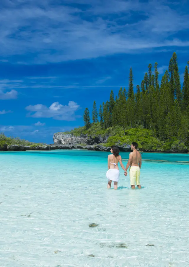 Un couple se tient main dans la main dans les eaux cristallines de la piscine naturelle de la baie d’Oro, à l’Île des Pins, entouré d’une végétation luxuriante et de pins colonnaires.