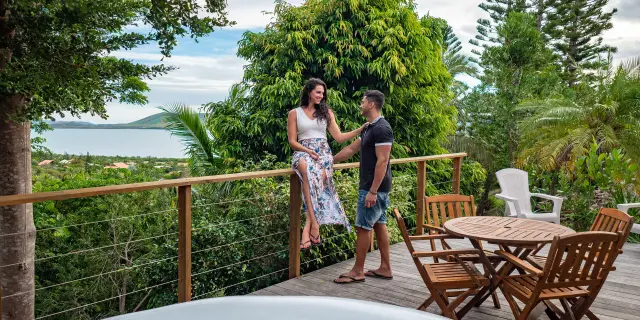 Couple sur la terrasse en bois du Oua-Ya Lodge à Boulouparis, avec bain de détente, mobilier extérieur, végétation tropicale et vue sur le lagon.