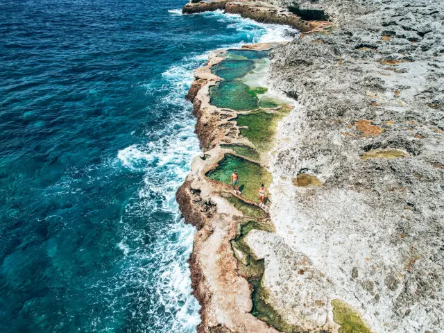 Vue aérienne des marmites du Cap des Pins à Lifou, avec des bassins naturels creusés dans la roche au bord de l’océan et deux personnes profitant de l’eau claire.