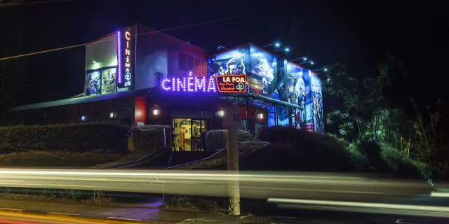 Illuminated façade of the Jean-Pierre Jeunet Cinema in La Foa, photographed at night with vehicle light trails in the foreground.