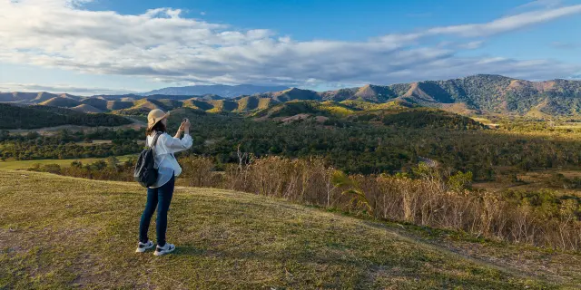 Une randonneuse photographie les collines du Domaine de Déva, entre reliefs verdoyants, ciel bleu et lumière de fin de journée.