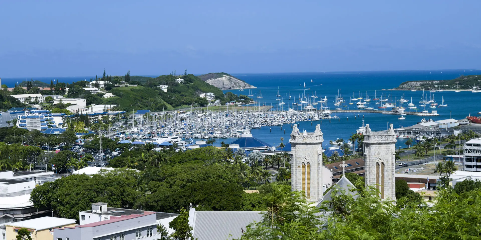 Vue panoramique sur la marina de Port Moselle à Nouméa, avec de nombreux voiliers, la cathédrale et le lagon en arrière-plan.