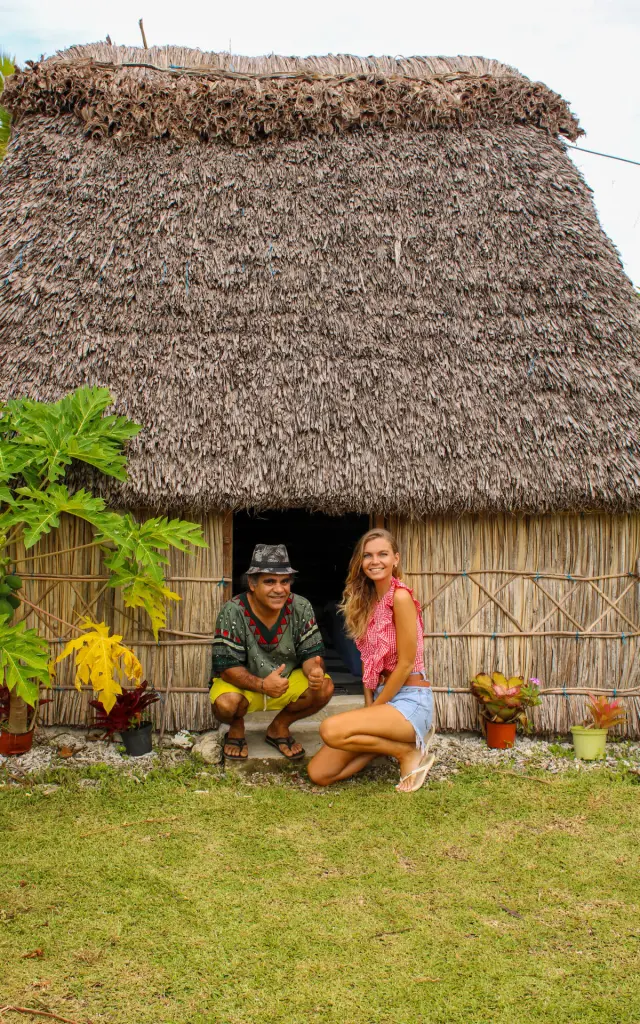 Traveller and host in front of a traditional hut at Gîte Chez Dom in Maré, with a woven palm-leaf roof and tropical vegetation.