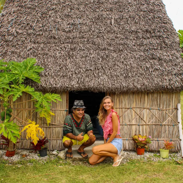 Voyageuse et hôte devant une case traditionnelle au gîte Chez Dom à Maré, avec toiture en feuilles de cocotier et végétation tropicale.