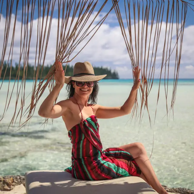 Une touriste néo-zélandaise assise au bord du lagon à l’Île des Pins, sous des feuilles de cocotier, face à l’eau turquoise.