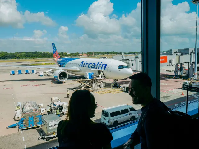 Un avion Aircalin stationné à l’aéroport international de Nouméa La Tontouta, vu depuis le terminal avec des voyageurs au premier plan.