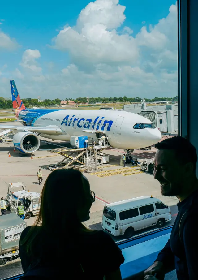 Un avion Aircalin stationné à l’aéroport international de Nouméa La Tontouta, vu depuis le terminal avec des voyageurs au premier plan.