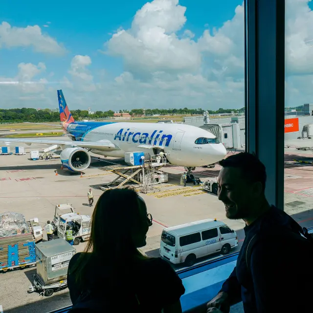 Un avion Aircalin stationné à l’aéroport international de Nouméa La Tontouta, vu depuis le terminal avec des voyageurs au premier plan.