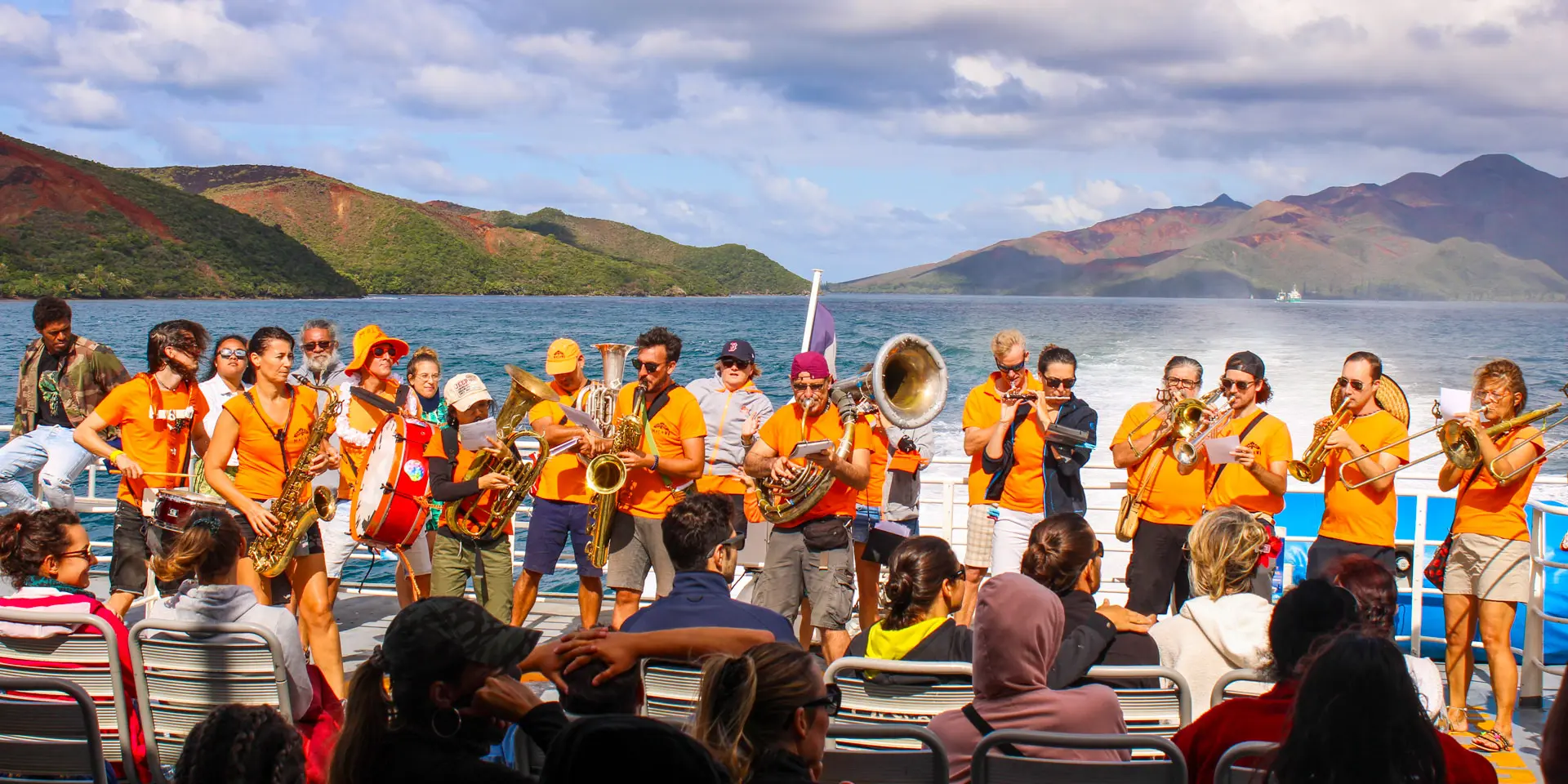 Un groupe de musiciens joue à bord du Betico devant les passagers, avec le lagon Sud et les montagnes en arrière-plan.