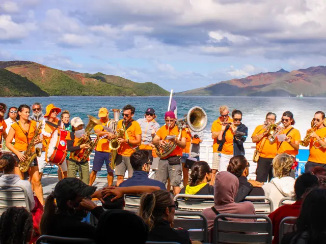 Un groupe de musiciens joue à bord du Betico devant les passagers, avec le lagon Sud et les montagnes en arrière-plan.