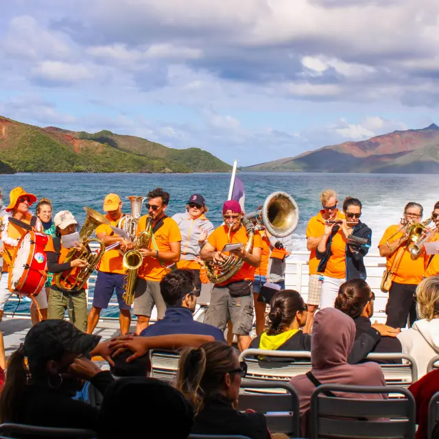 Un groupe de musiciens joue à bord du Betico devant les passagers, avec le lagon Sud et les montagnes en arrière-plan.