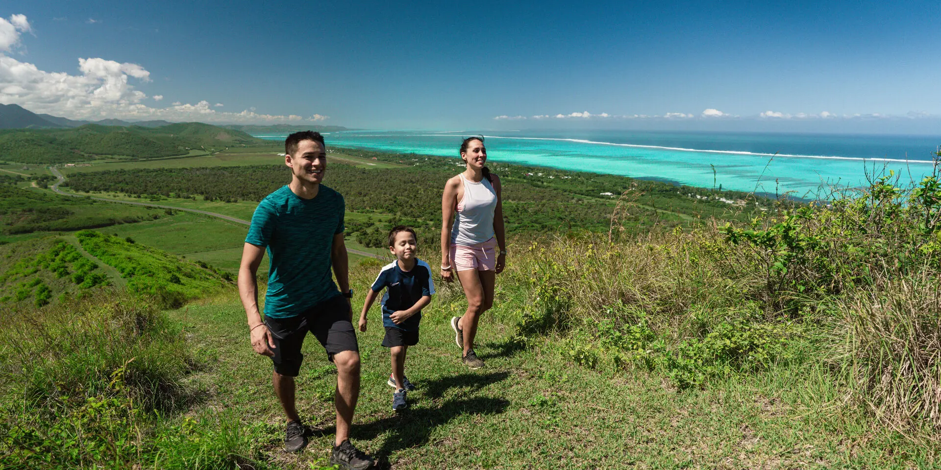 Une famille en randonnée sur un sentier du Domaine de Déva, avec le lagon turquoise en arrière-plan, en Nouvelle-Calédonie.