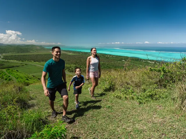 Une famille en randonnée sur un sentier du Domaine de Déva, avec le lagon turquoise en arrière-plan, en Nouvelle-Calédonie.