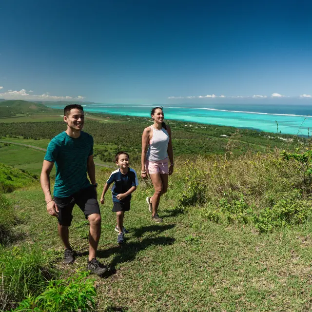 Une famille en randonnée sur un sentier du Domaine de Déva, avec le lagon turquoise en arrière-plan, en Nouvelle-Calédonie.