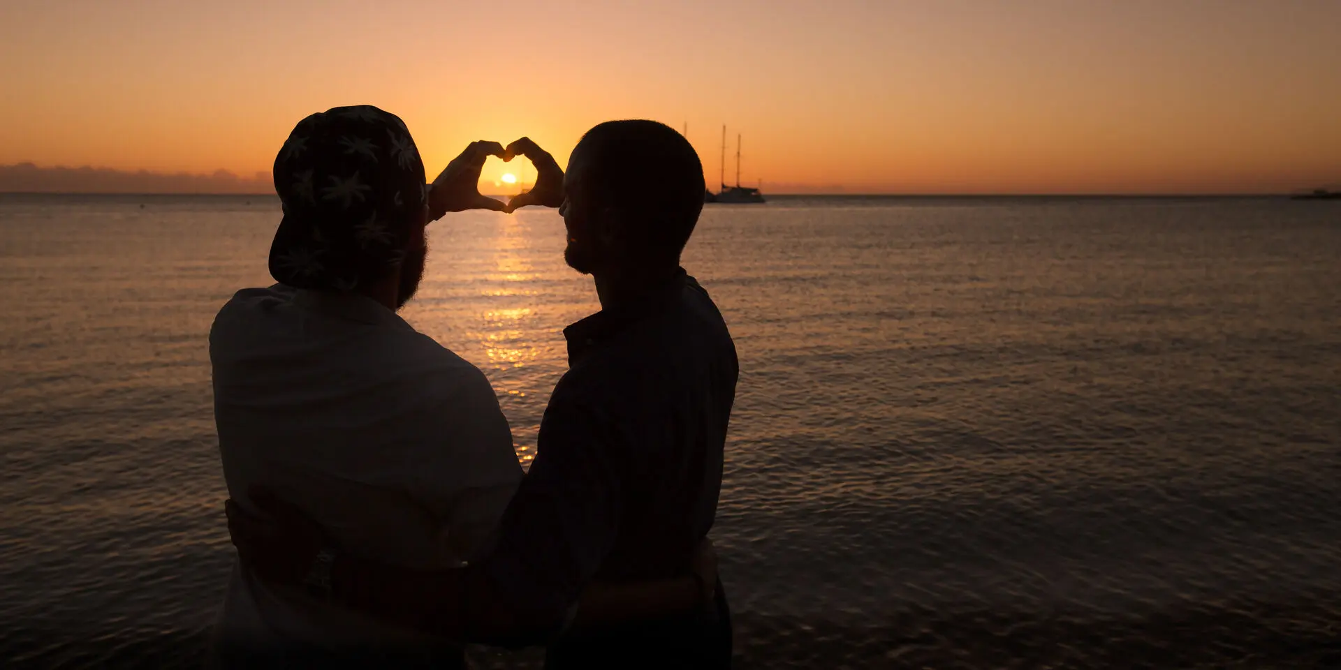 Silhouette d’un couple sur la plage de la Baie des Citrons à Nouméa, formant un cœur avec les mains devant le soleil couchant.