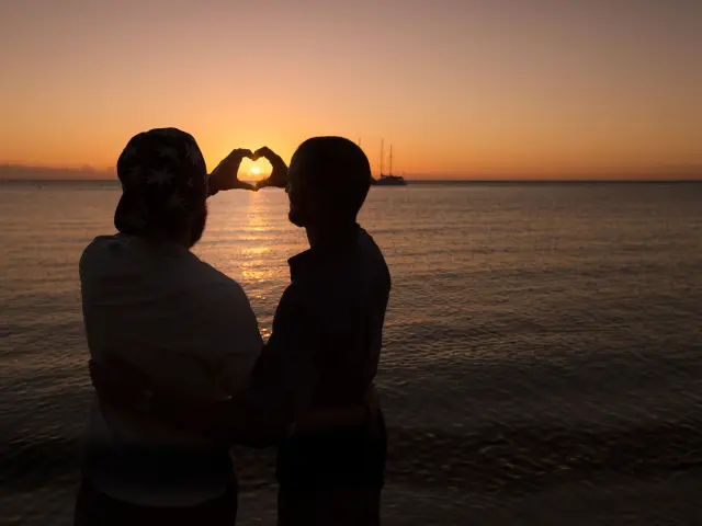 Silhouette d’un couple sur la plage de la Baie des Citrons à Nouméa, formant un cœur avec les mains devant le soleil couchant.