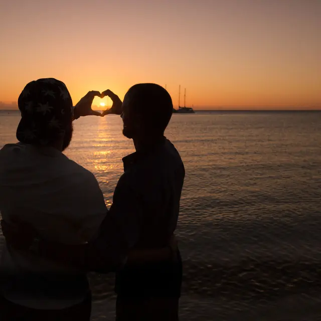 Silhouette d’un couple sur la plage de la Baie des Citrons à Nouméa, formant un cœur avec les mains devant le soleil couchant.