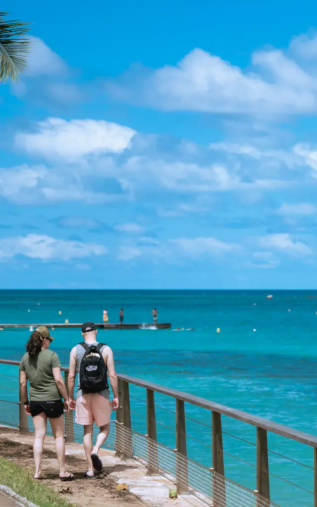 Deux touristes marchent sur la promenade de l’Anse Vata à Nouméa, face au lagon turquoise.