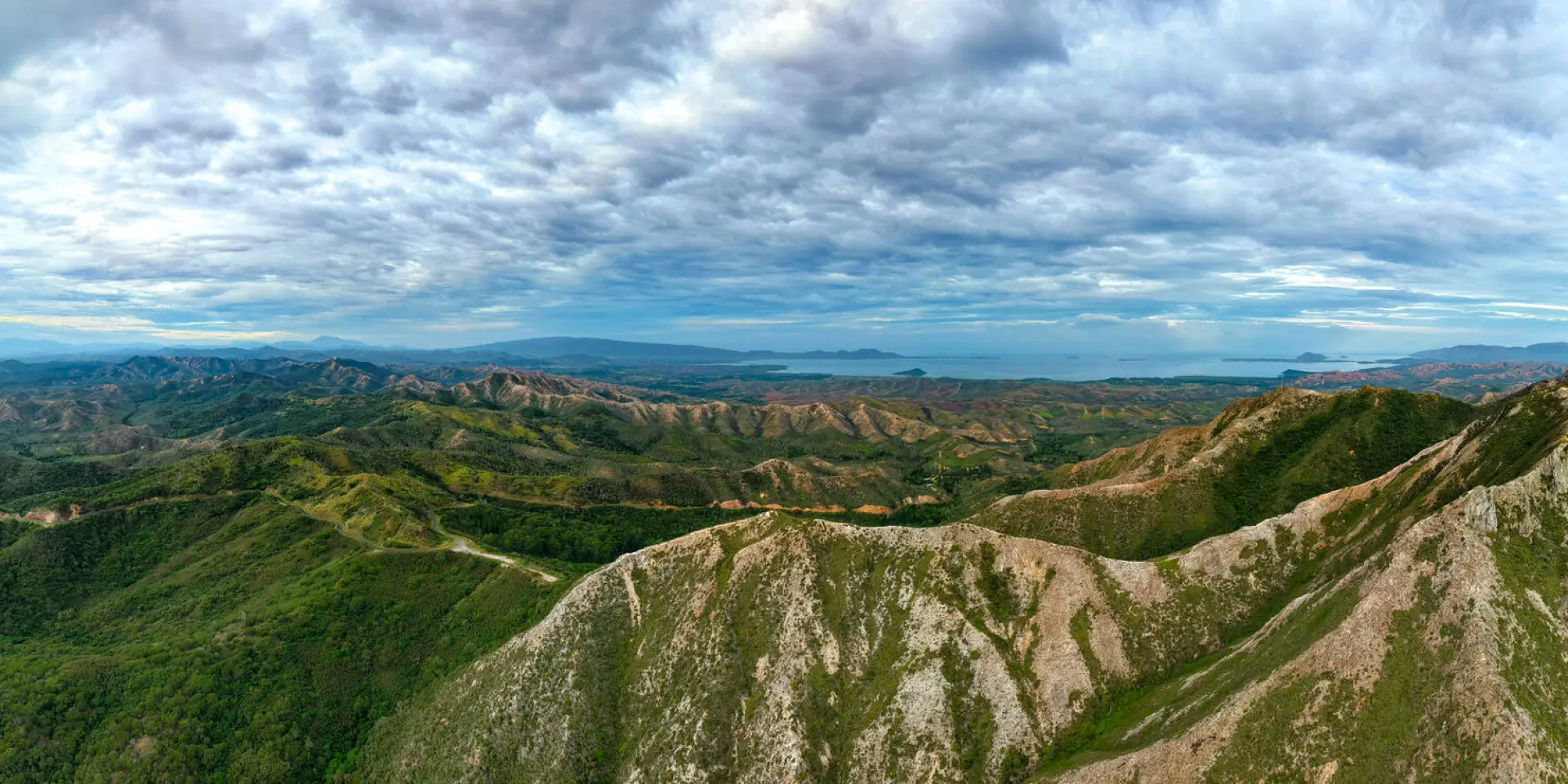 Panorama depuis le col d’Arama à Poum, avec les crêtes verdoyantes au premier plan et la mer au loin sous un ciel nuageux.