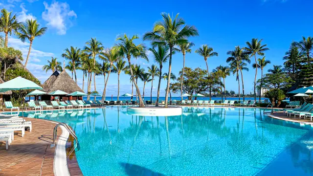 Piscine du Domaine Nouméa bordée de palmiers, avec transats et vue sur la mer.