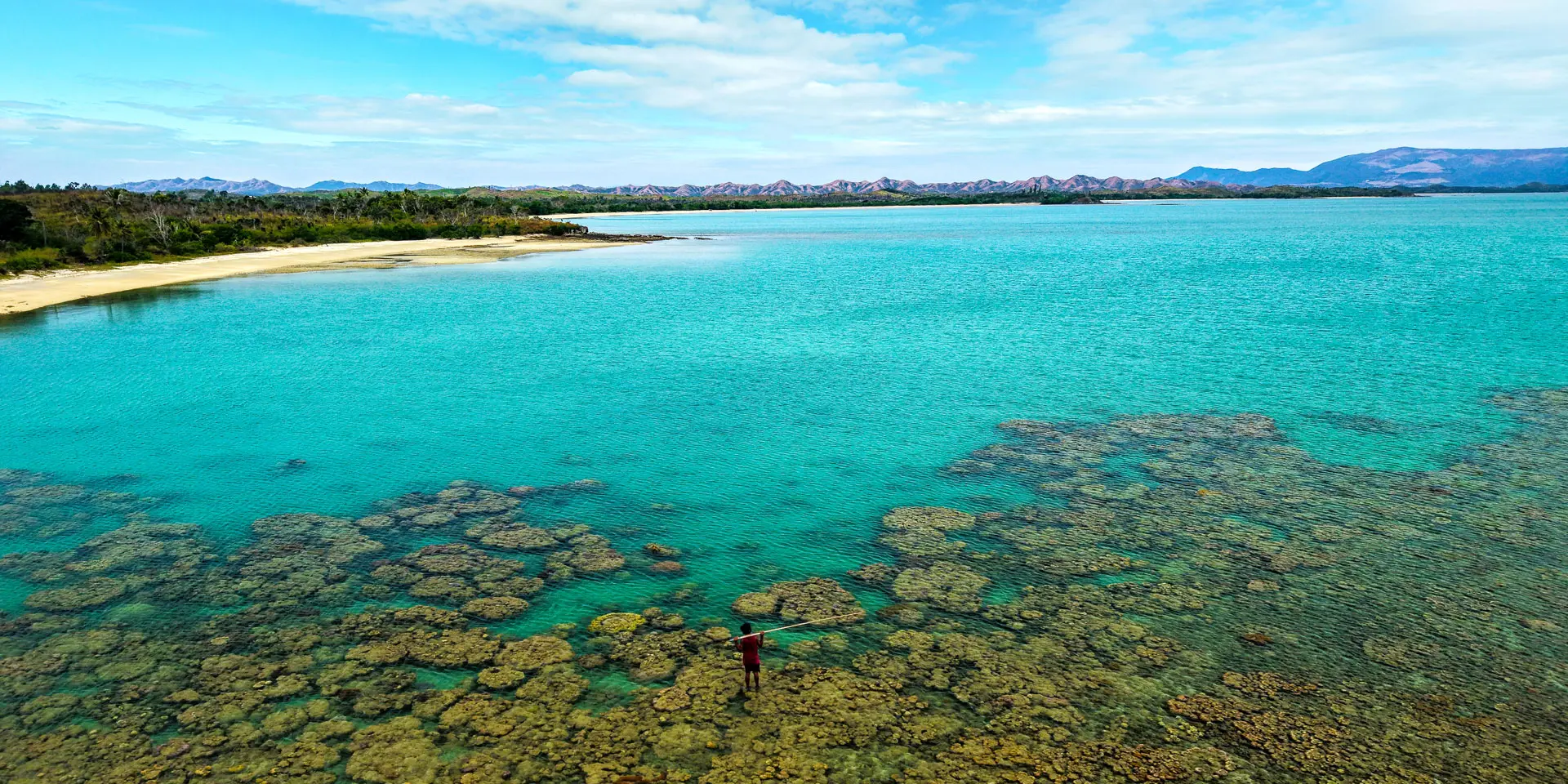 Vue aérienne de la plage de Nénon à Poum, avec lagon turquoise, patates de corail et pêcheur debout sur le récif.
