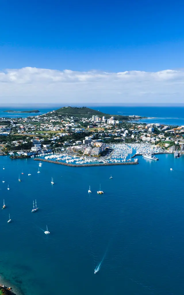 Vue aérienne de Nouméa : la ville, ses baies et de nombreux voiliers au mouillage dans un lagon bleu.
