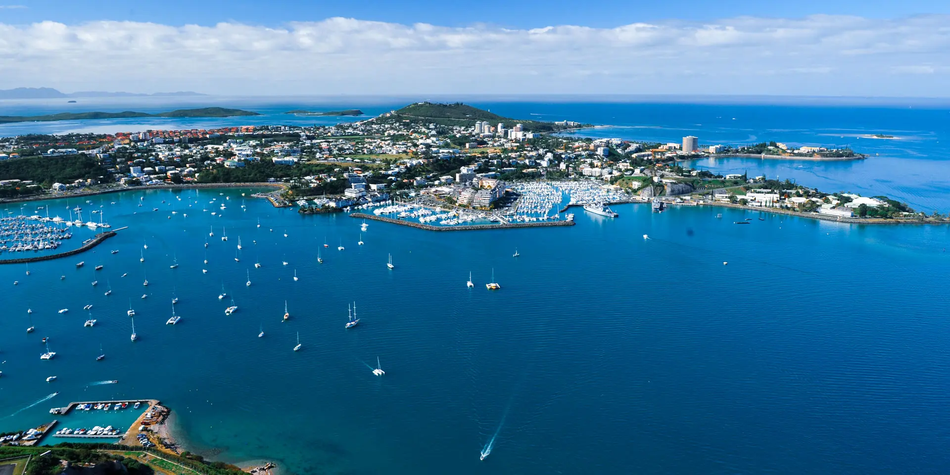 Vue aérienne de Nouméa : la ville, ses baies et de nombreux voiliers au mouillage dans un lagon bleu.