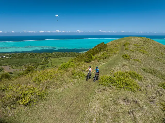 Deux cyclistes roulent sur un sentier du Domaine de Déva à Bourail, avec le lagon turquoise et la barrière de corail en arrière-plan.