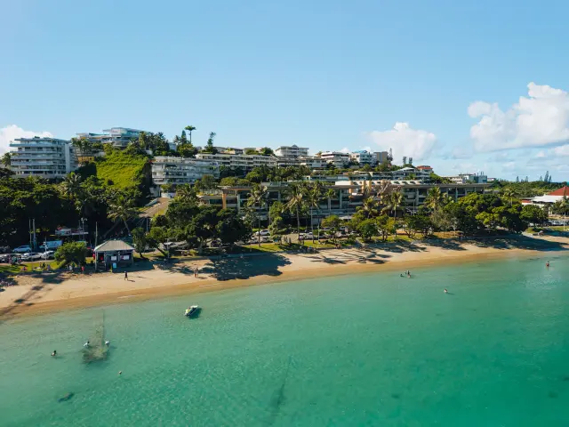 Vue aérienne de la Baie des Citrons à Nouméa, avec sa plage de sable, son eau turquoise, quelques baigneurs et les immeubles en bord de mer.