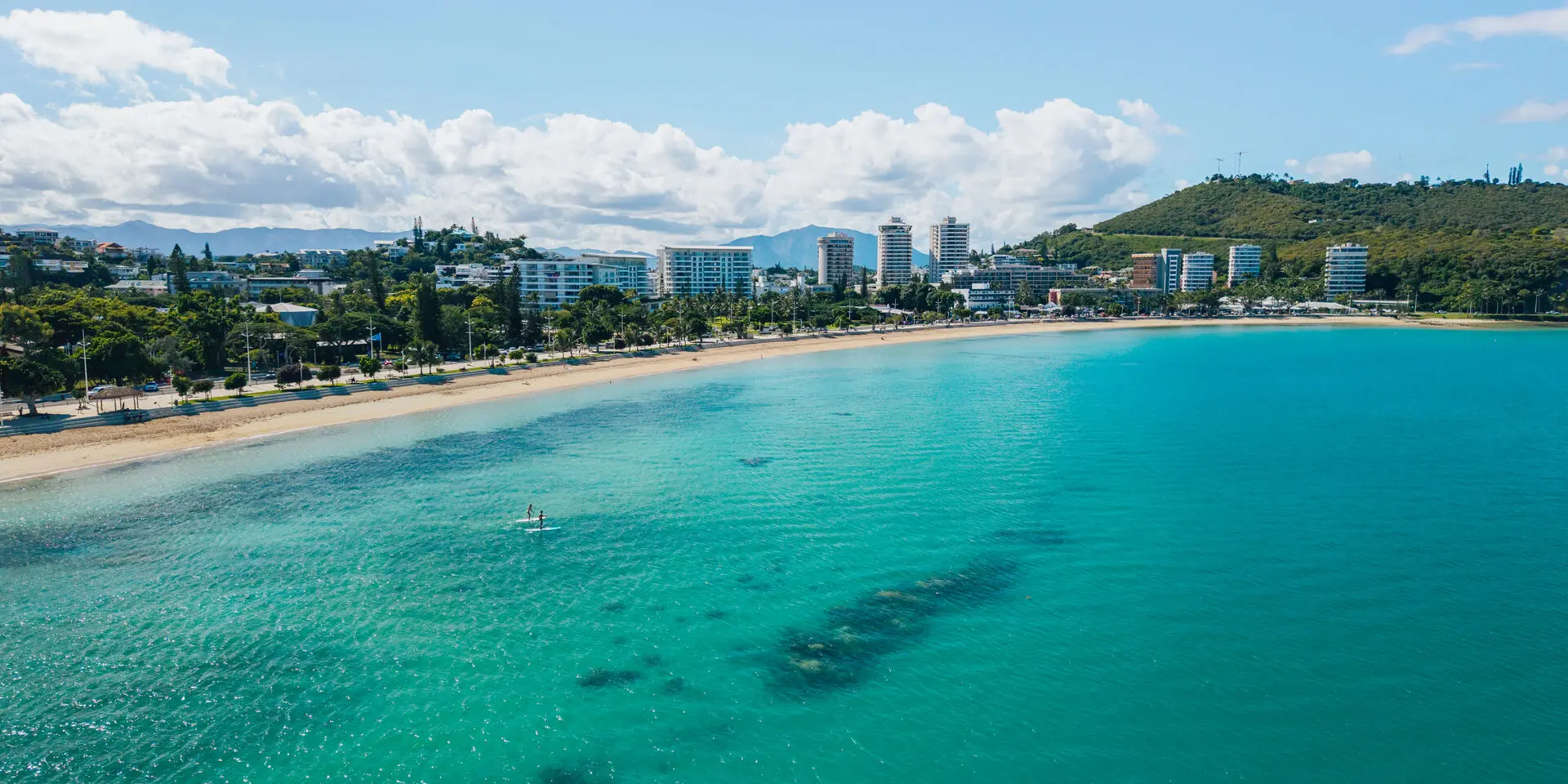 Aerial view of Anse Vata Bay in Nouméa, featuring its long sandy beach, turquoise water, a few people on the water and the waterfront buildings.