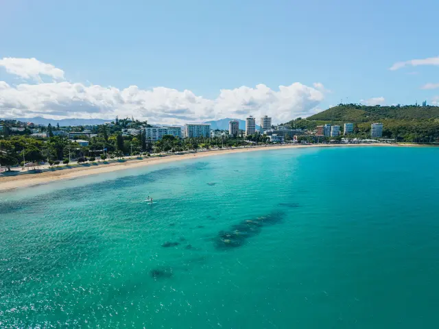 Aerial view of Anse Vata Bay in Nouméa, featuring its long sandy beach, turquoise water, a few people on the water and the waterfront buildings.