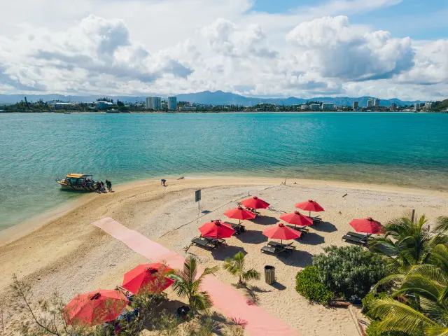 Vue depuis l’îlot Canard à Nouméa, avec sa plage de sable bordée de parasols rouges, un bateau accosté au rivage et la capitale en arrière-plan.