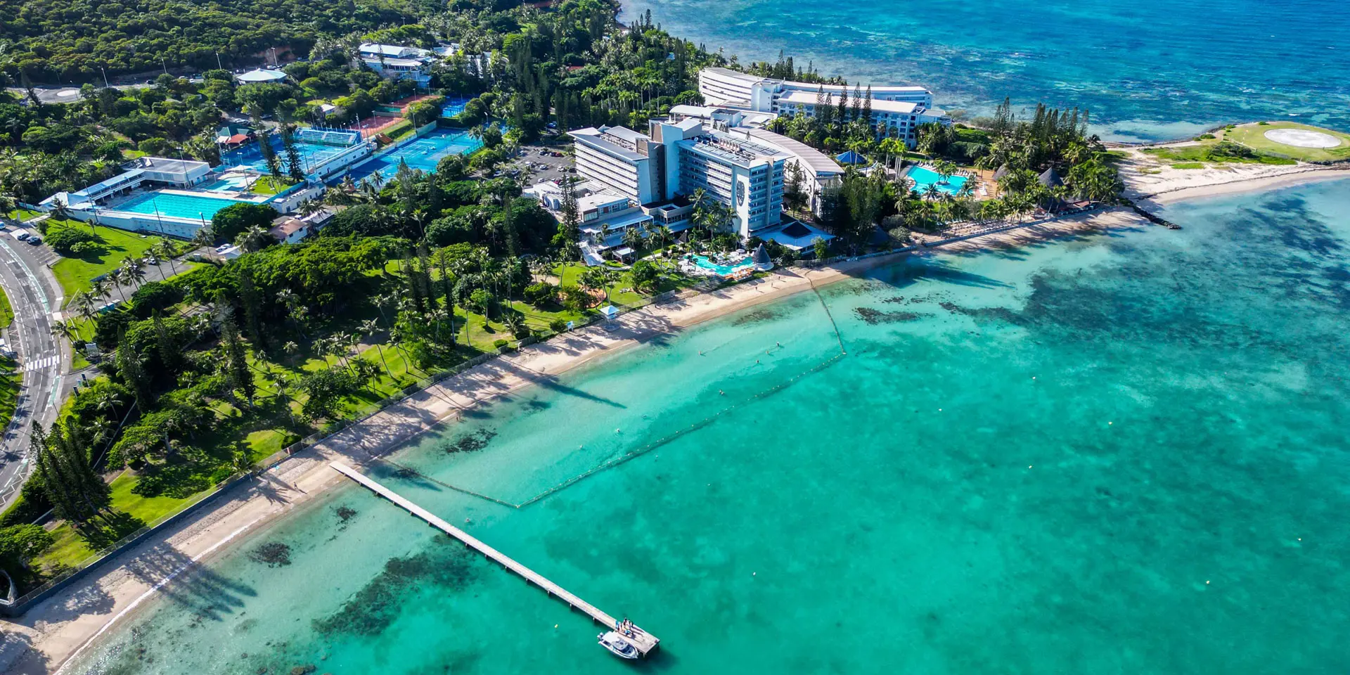 Aerial view of Château Royal Beach Resort & Spa in Nouméa, set beside the lagoon with a beach, swimming pool and jetty facing turquoise waters.