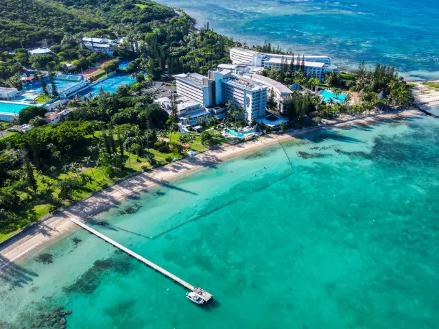Aerial view of Château Royal Beach Resort & Spa in Nouméa, set beside the lagoon with a beach, swimming pool and jetty facing turquoise waters.