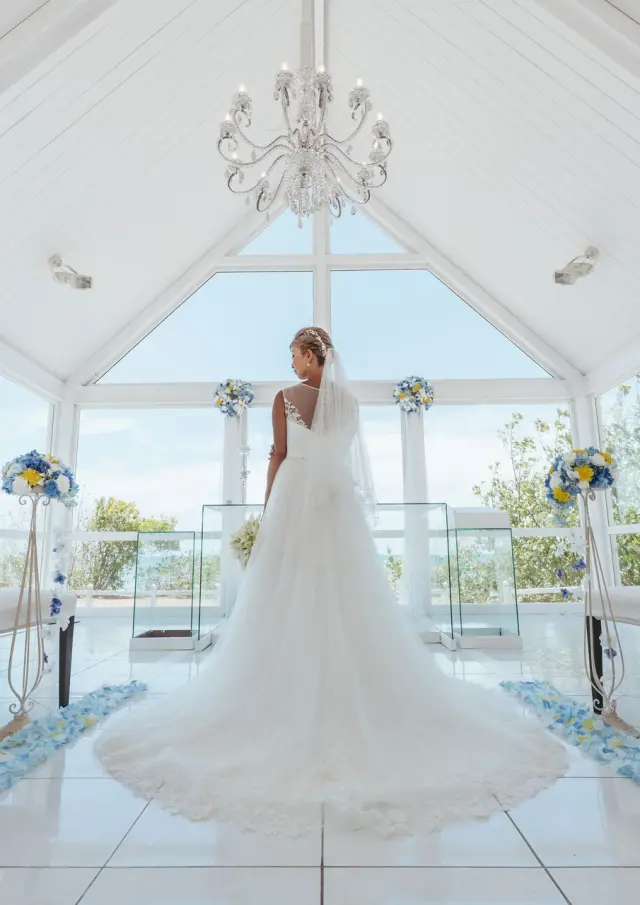 A bride in a white gown stands in a bright hotel chapel in Nouméa, decorated with white, blue and yellow flowers.
