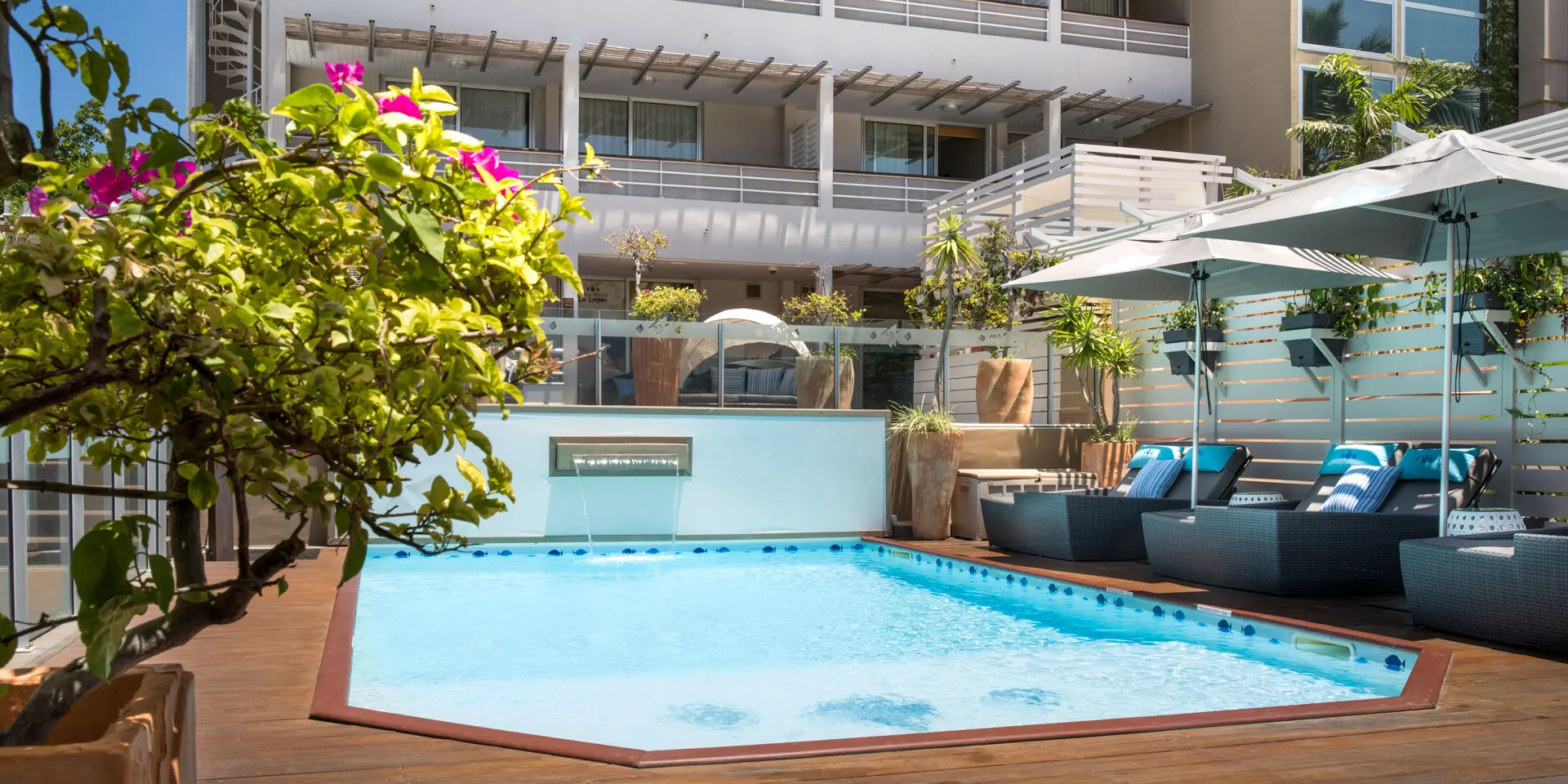 Swimming pool at Hôtel Le Lagon in Nouméa, surrounded by sun loungers, umbrellas and tropical greenery in an urban setting.