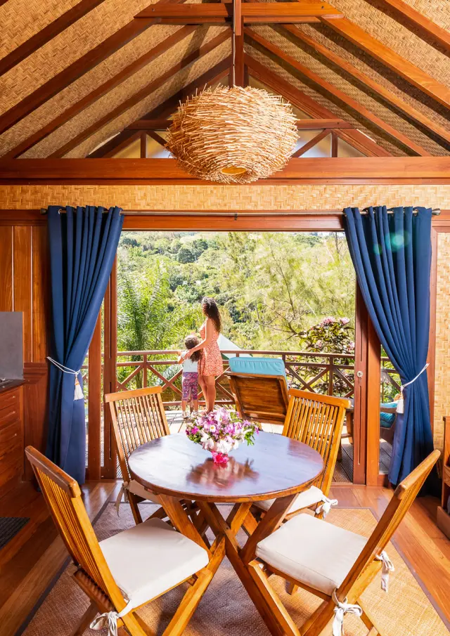 Vue intérieure d’un bungalow en bois à l’Oasis de Tendéa à Farino, avec coin cuisine, table, banquette et terrasse ouverte sur la nature.