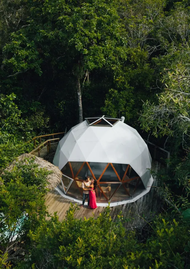 Vue aérienne d’un dôme des Bulles de Farino, niché au cœur de la végétation, avec un couple sur la terrasse en bois.