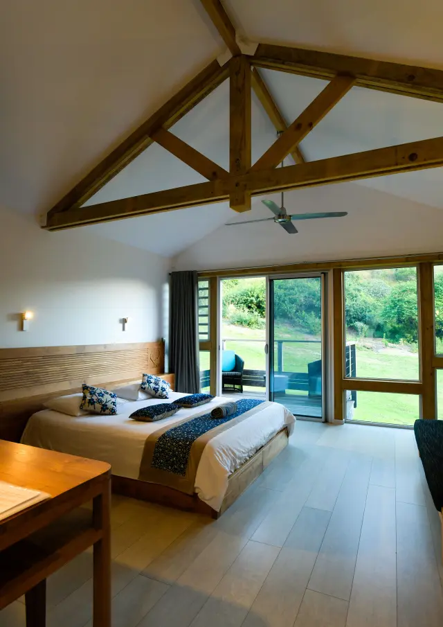 Intérieur d’une chambre lumineuse au Betikuré Parc Lodge à Bourail, avec lit double, charpente apparente et baie vitrée ouverte sur la verdure.