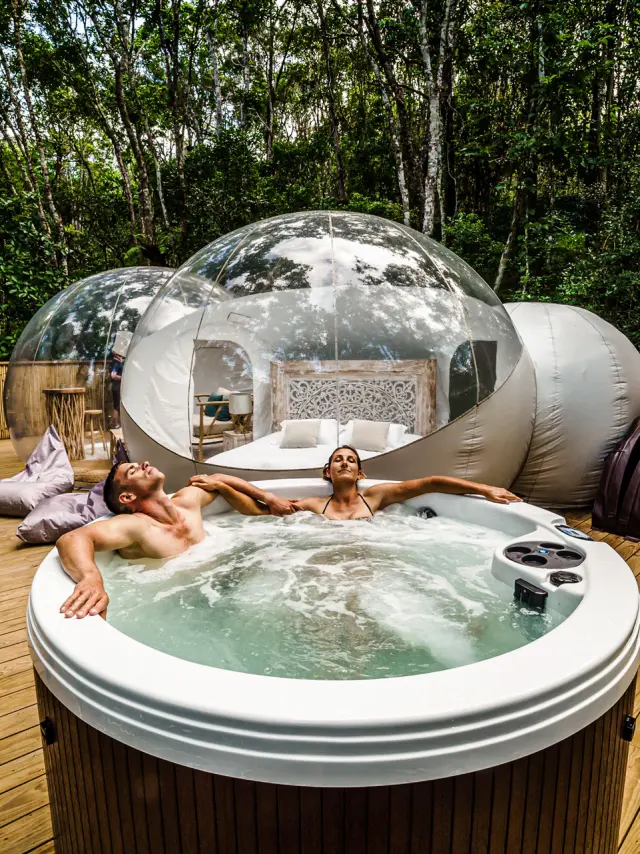 Un couple se détend dans un jacuzzi sur une terrasse en bois, devant une bulle transparente installée en pleine forêt aux Bulles de Farino.