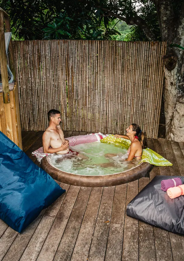 Couple se détendant dans un bain à remous sur la terrasse en bois du Lodge Terre de Soleil à Farino, entourée d’arbres et de végétation tropicale.