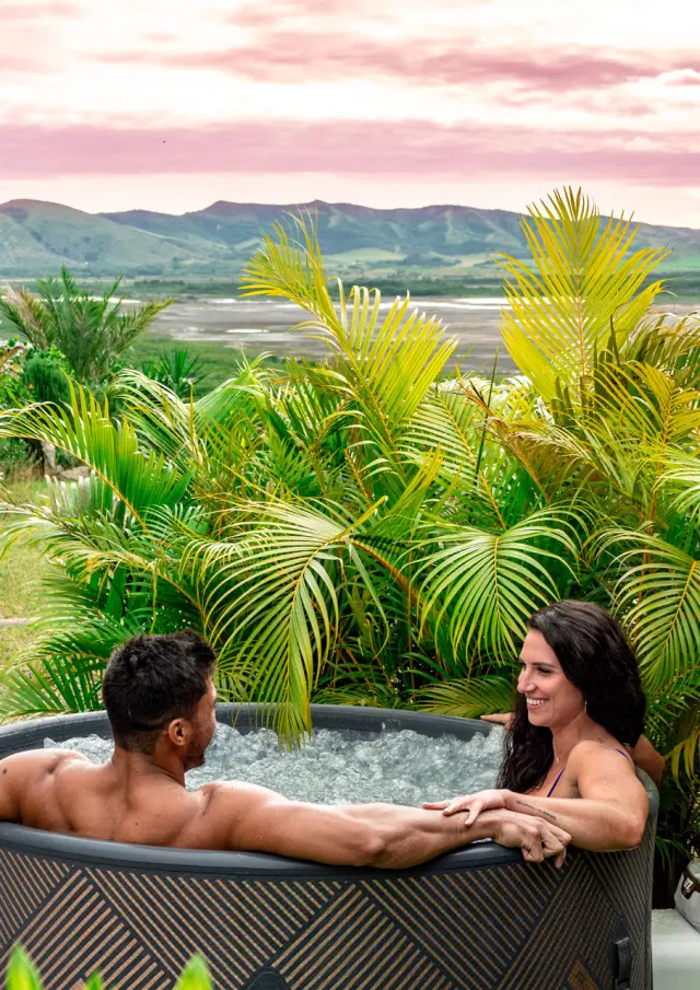 Couple se relaxant dans un bain extérieur au Oua-Ya Lodge à Boulouparis, entouré de palmiers avec vue sur les collines et le lagon au coucher du soleil.