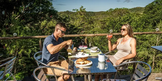 Couple prenant le petit-déjeuner sur la terrasse de l’espace restauration du Lodge Terre de Soleil à Farino, avec vue sur la végétation et les collines.